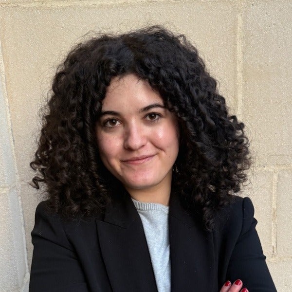 Sarah Aaron, dark curly hair, warm smile, black blazer over light blue sweater, beige background