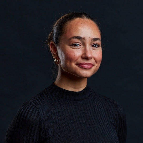 Sarolta Lukovszky, dark hair pulled back, warm smile, gold hoop earrings, black ribbed sweater, dark background