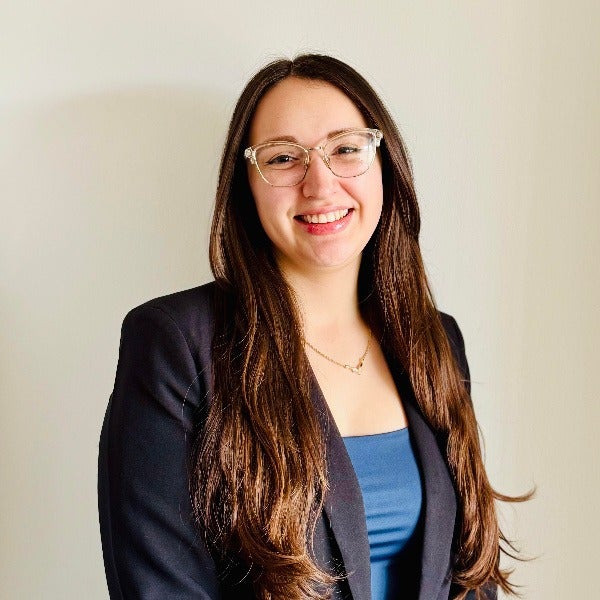 Taylor Elliott, long brown hair, glasses, warm smile, blue top, dark blazer, light background