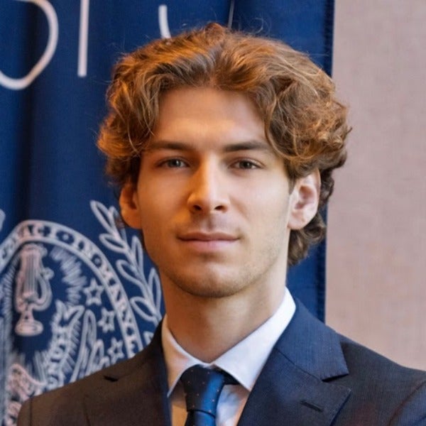 Theo Bogaty, wavy brown hair, light skin, subtle smile, dark suit with blue tie, blue background