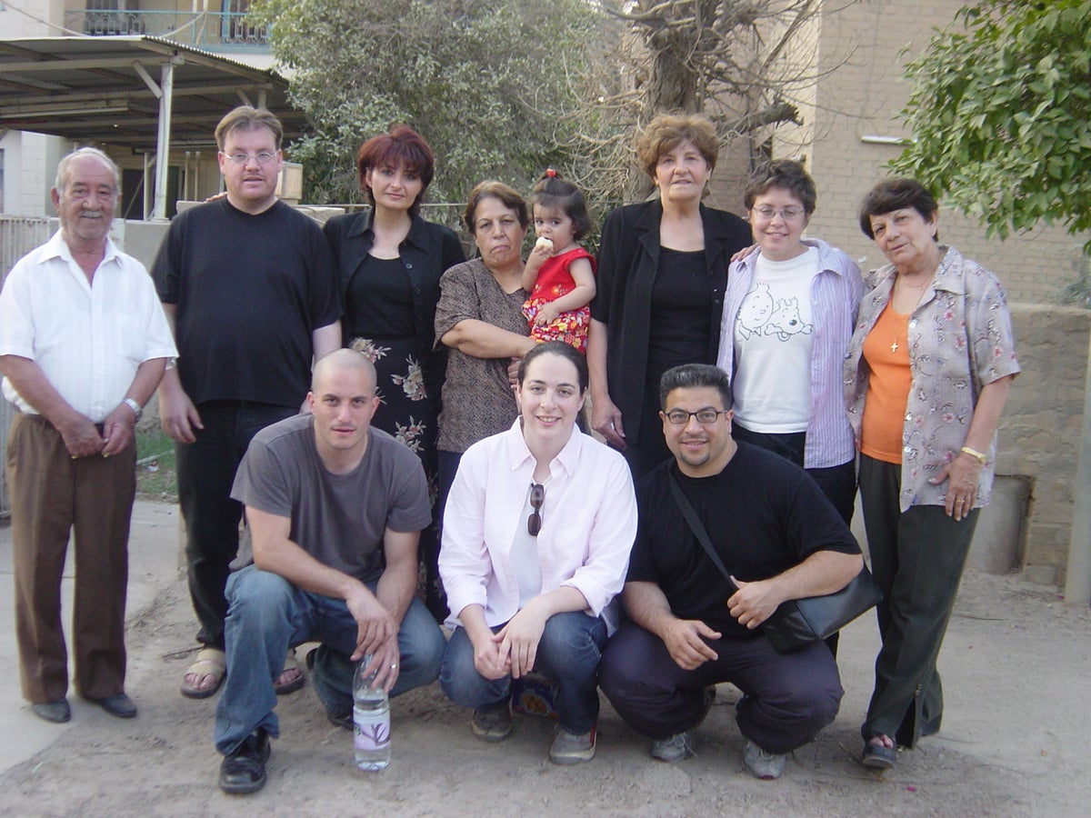 A group of eleven people, including adults and a child, is posing outdoors in a residential area with trees and buildings in the background. The group is casually dressed and some are standing while others are crouching in the front.