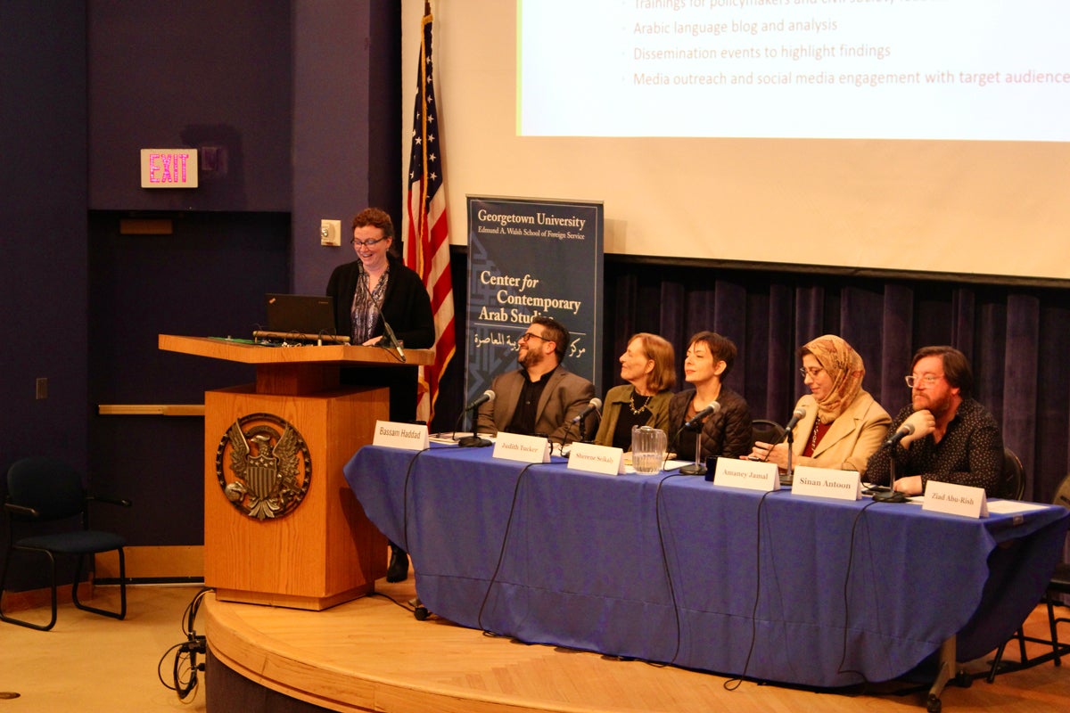 A panel discussion at Georgetown University’s Center for Contemporary Arab Studies. Six individuals are seated at a table with microphones, and one person stands at a podium speaking. A screen displays some text in the background, and a banner for the Center is placed between the podium and the table. An American flag is visible behind the podium.