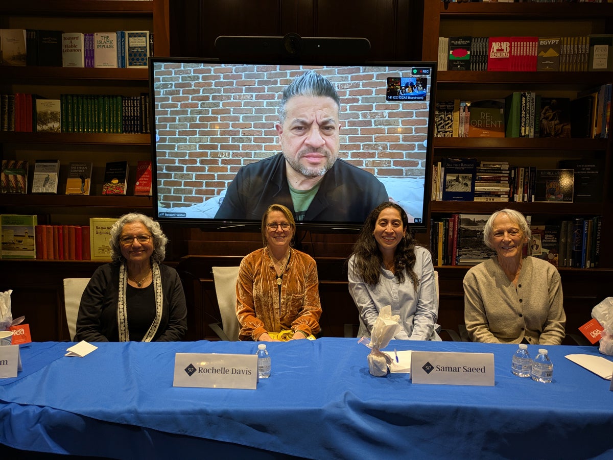 Four individuals are seated at a table with a blue tablecloth, featuring name cards for Rochelle Davis and Samar Saeed. Behind them is a large screen displaying a person in front of a brick wall. The background shows shelves filled with books.