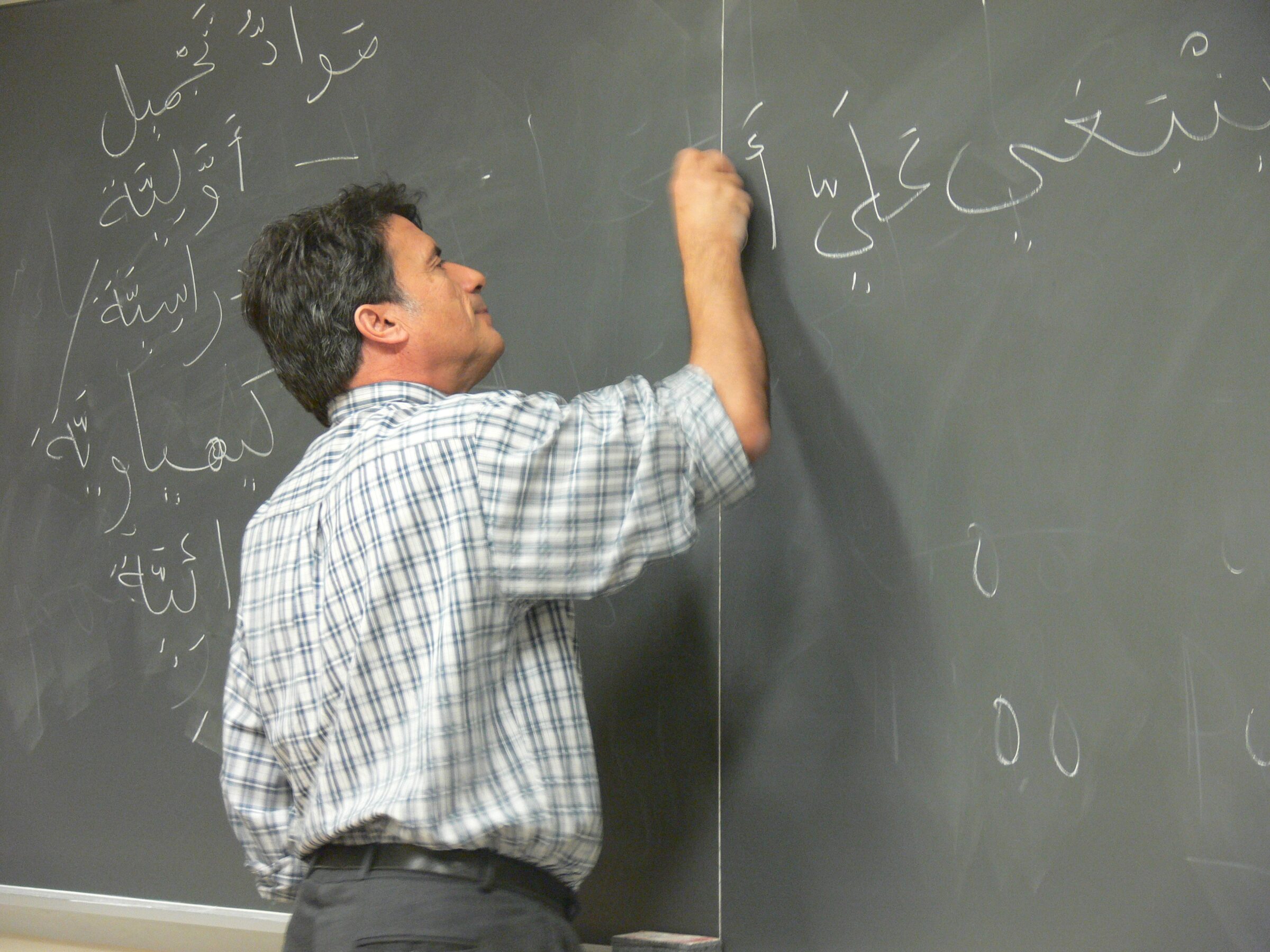 A man in a checked shirt with sleeves rolled up writes on a blackboard with chalk.