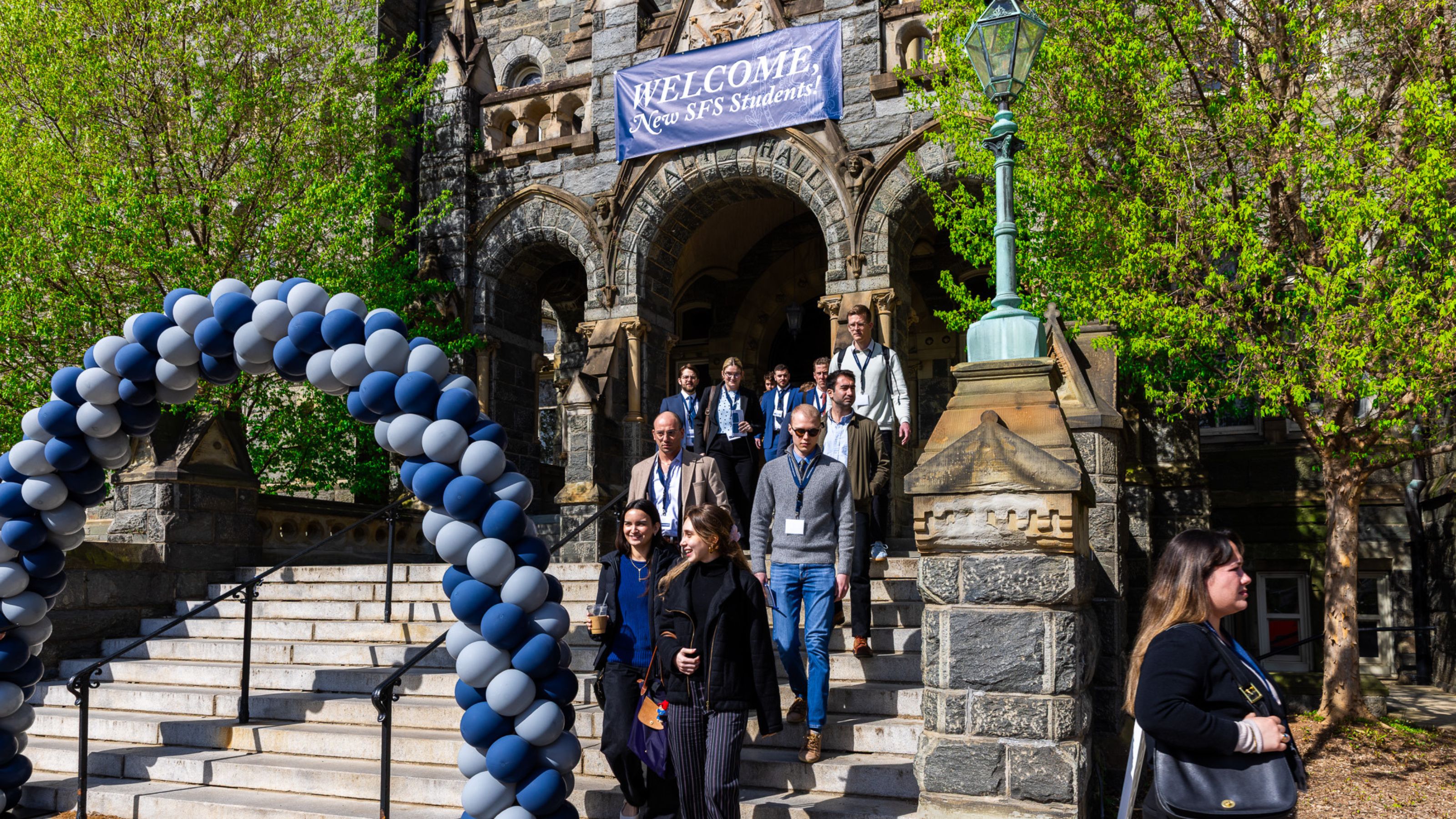 A group of people walking down the steps of a historic building at Georgetown University. A "Welcome SFS Students" banner is displayed above them. Blue and white balloon arches are positioned on the stairs. Lush greenery surrounds the scene.