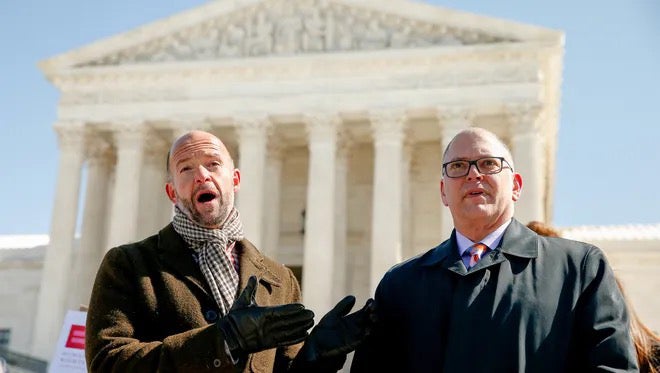 Two individuals stand in front of the United States Supreme Court building, having a conversation. Both are dressed in formal winter attire. The building's columns and pediment are visible in the background.
