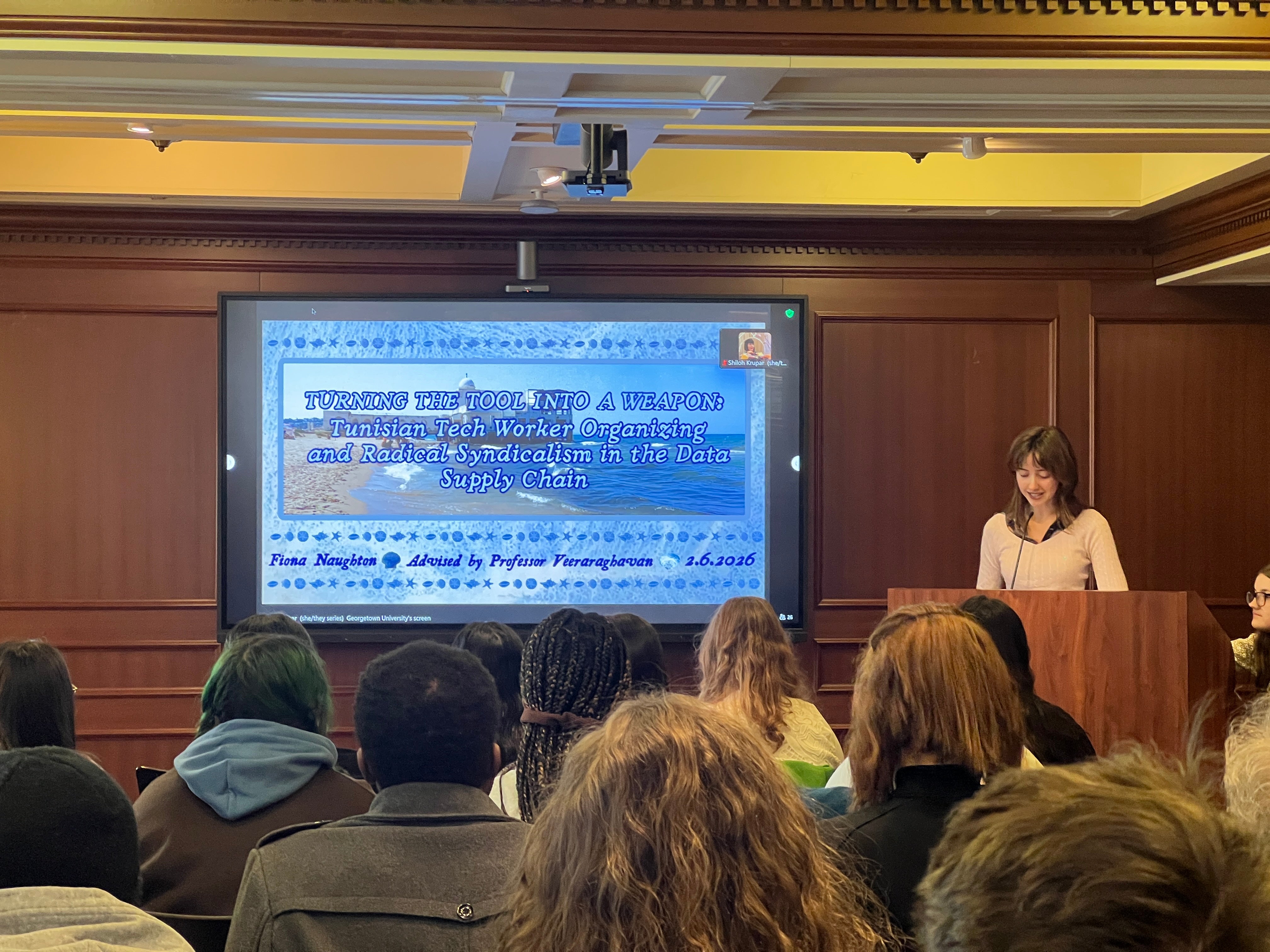 A young woman stands at a podium giving a presentation on a series of slides to a full audience.