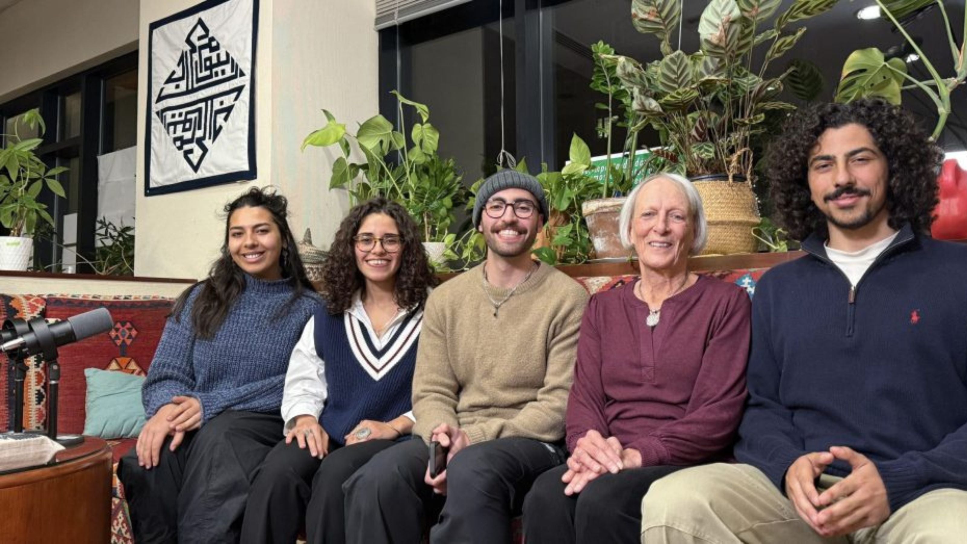 A group of five people sitting on a couch, smiling at the camera. Behind them is a wall hanging with Arabic calligraphy, and there are various green plants around. A microphone is visible on the table in front of them.