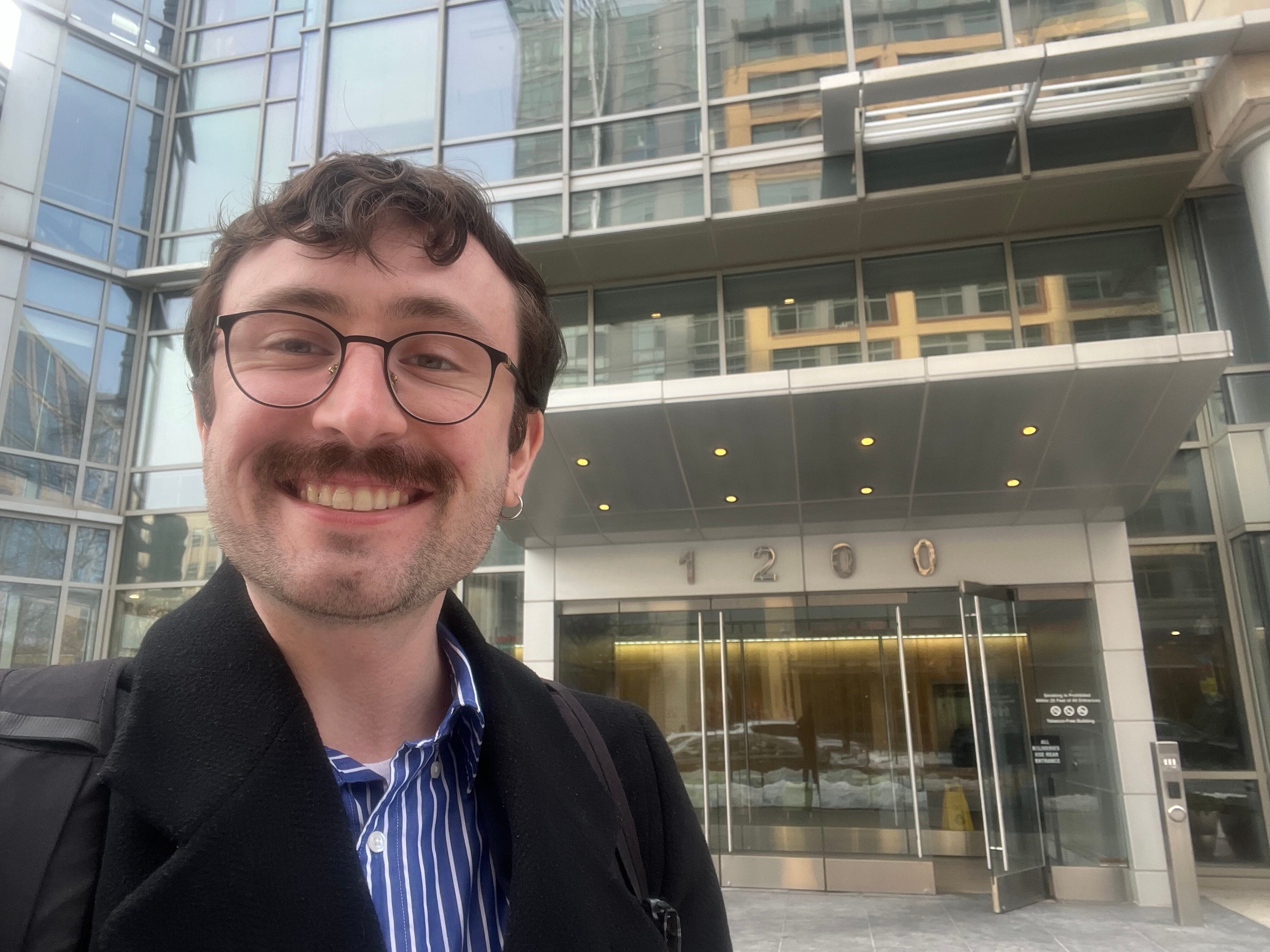 An individual with brown hair and glasses taking a selfie in front of a glass building.