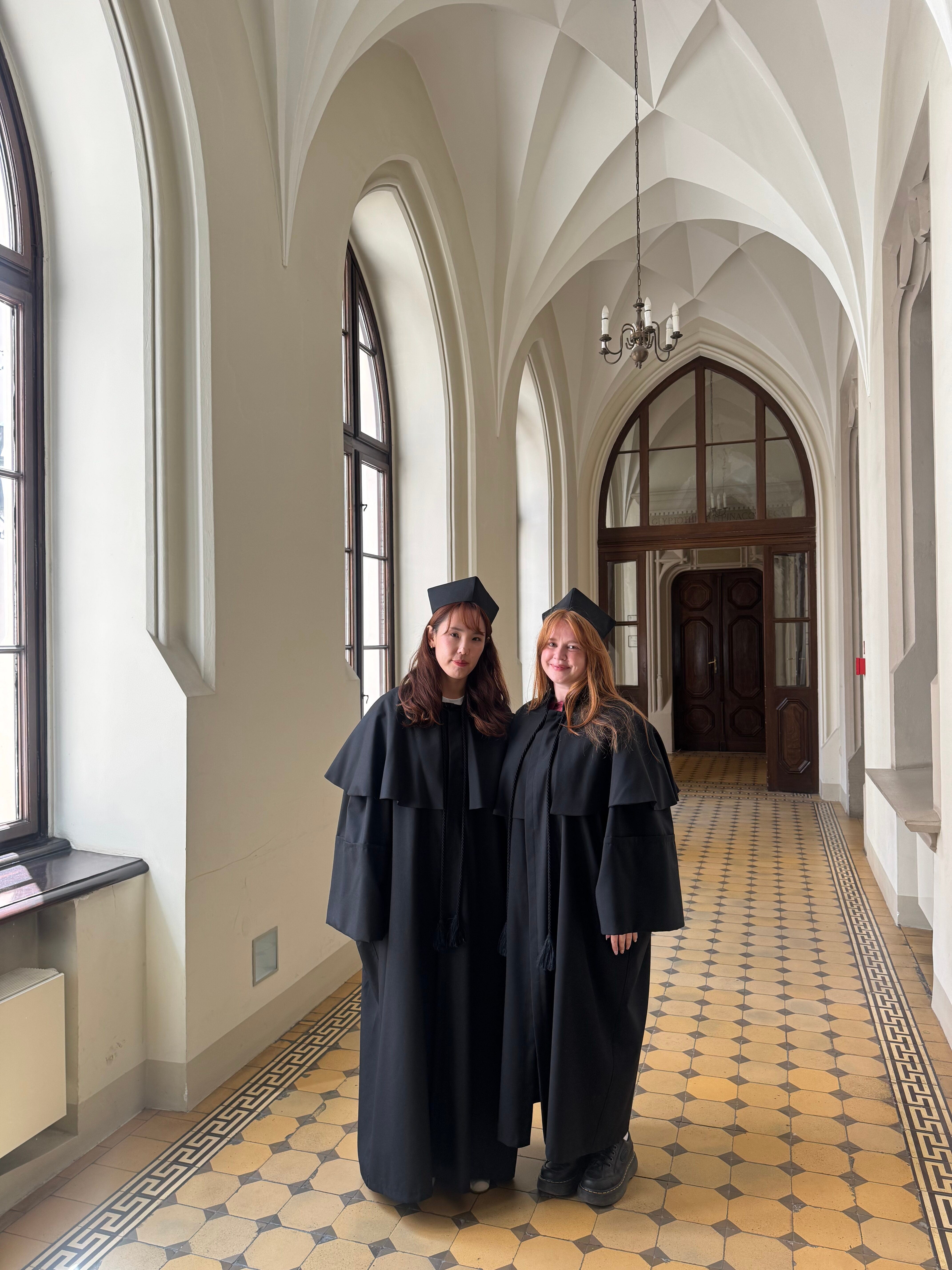 Two people in graduation attire stand side by side in a hallway with arched windows and a patterned tile floor. They are wearing black academic gowns and caps.