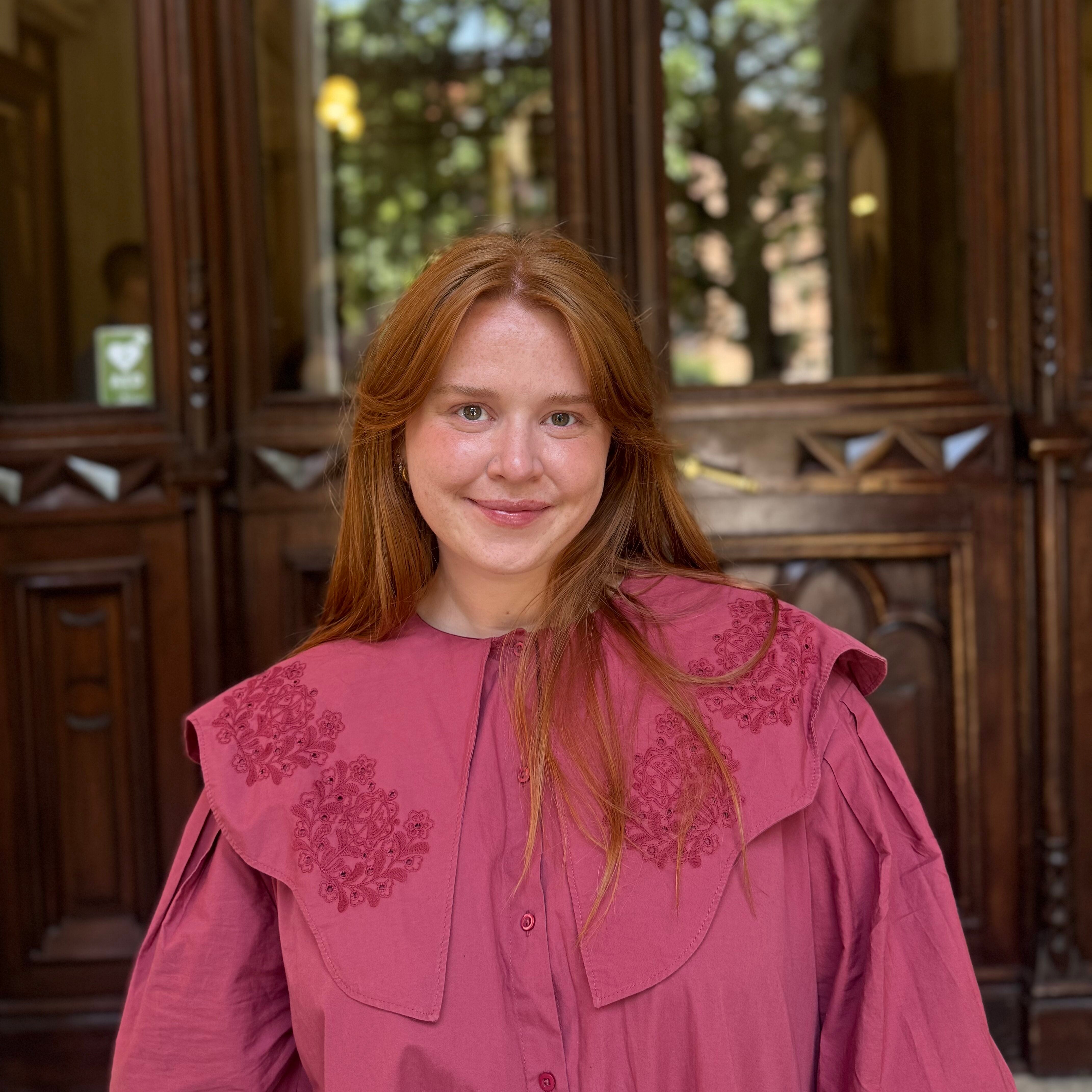 An individual with long red hair wearing a pink dress stands in front of an ornate brown wooden door.