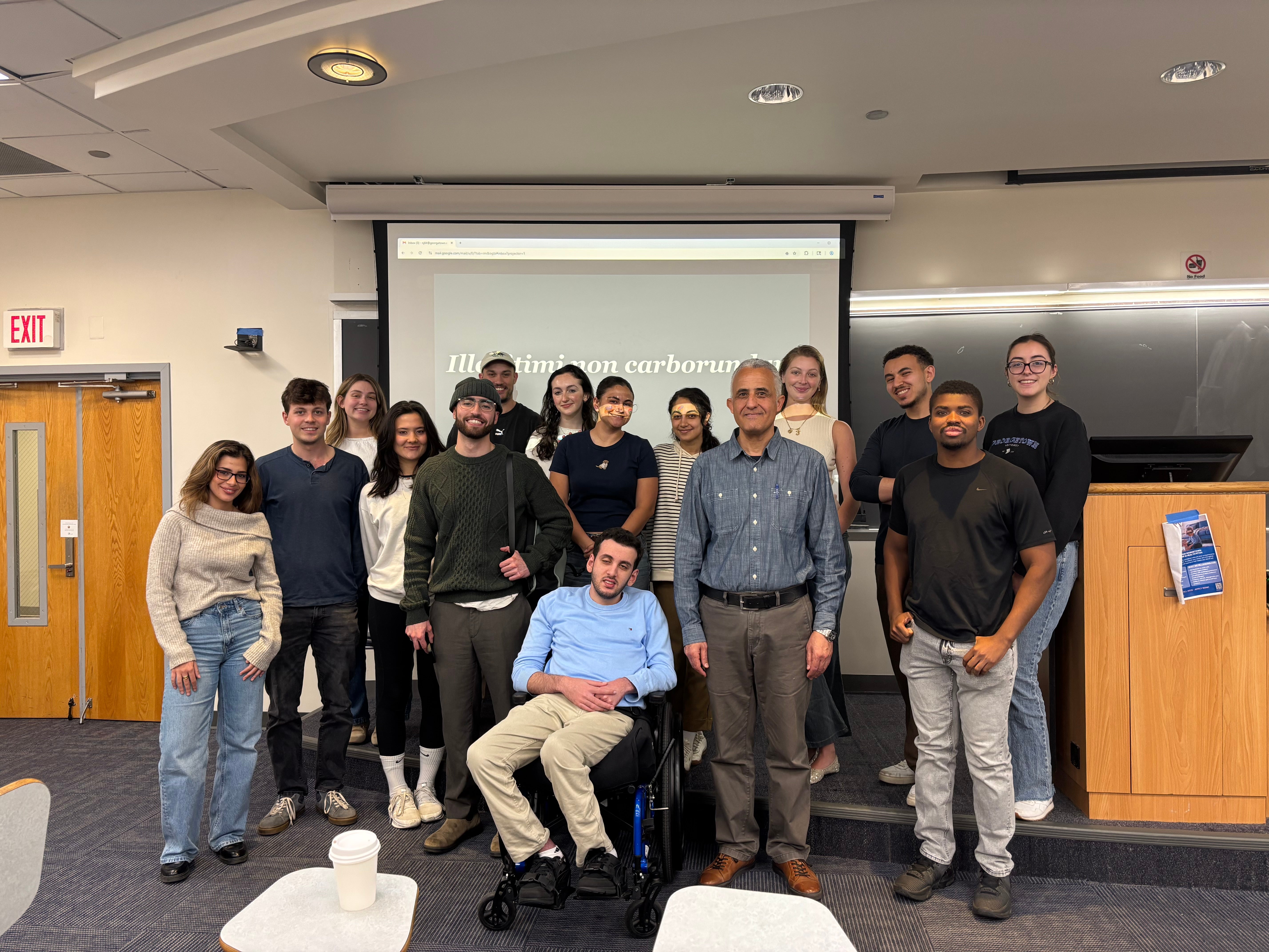 Fourteen people stand at the front of a classroom in two rows and smile.