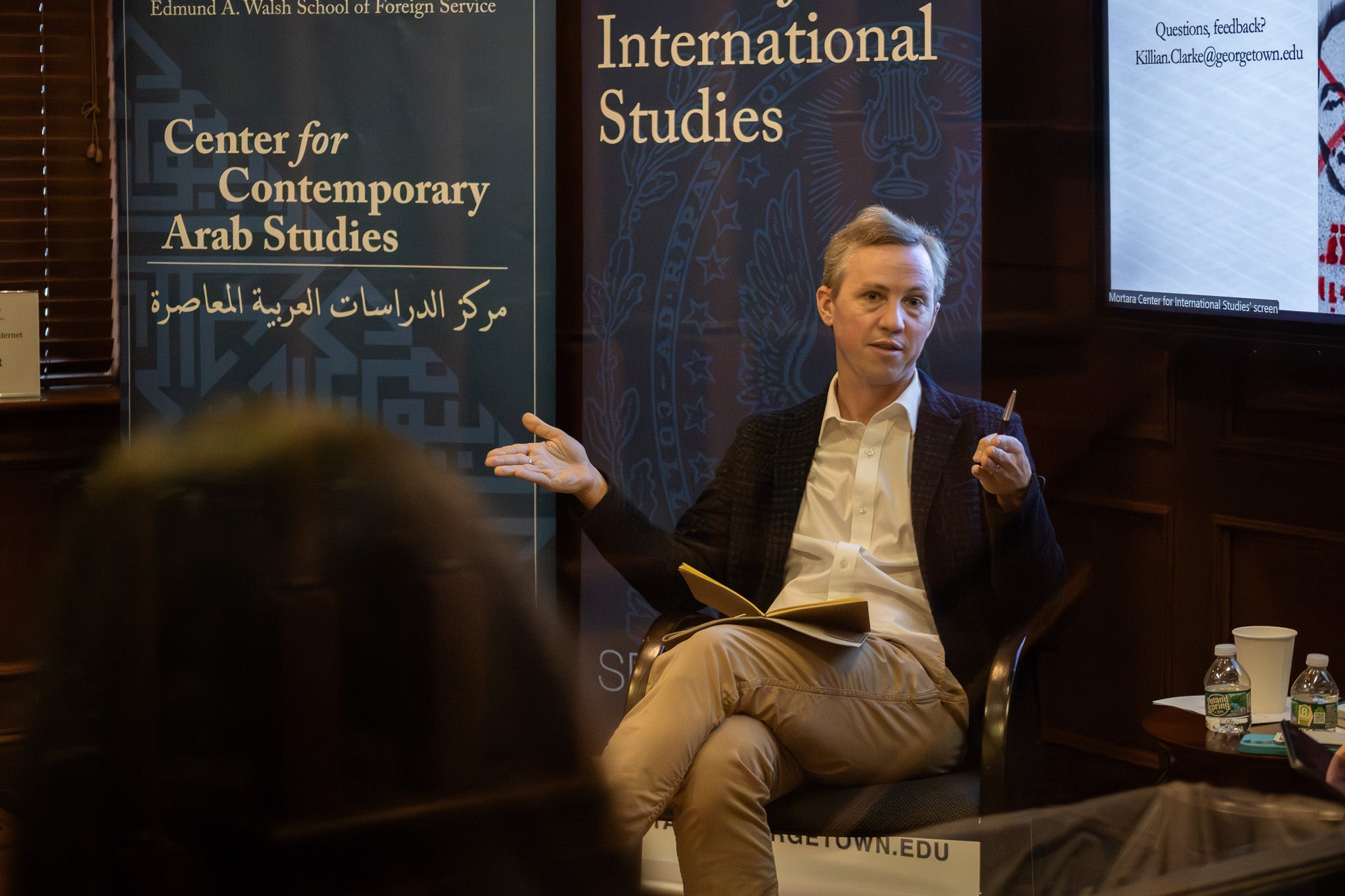 White man with dark blue jacket and white shirt sits in a chair with a book on his lap
