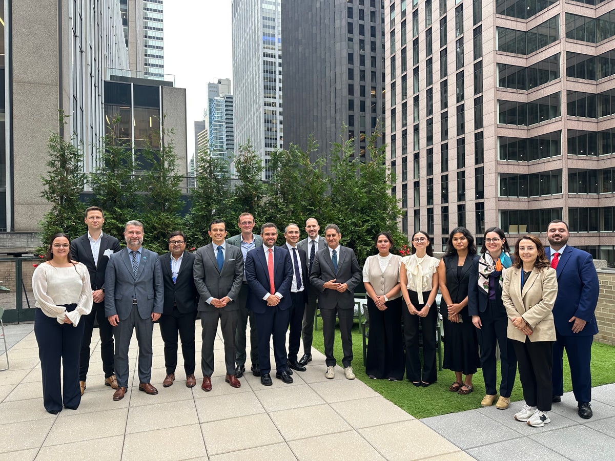 A group of individuals stands together outdoors on a city rooftop surrounded by tall buildings. Some people are dressed in formal attire, with a mix of suits and business wear. The setting appears urban with greenery in the background.