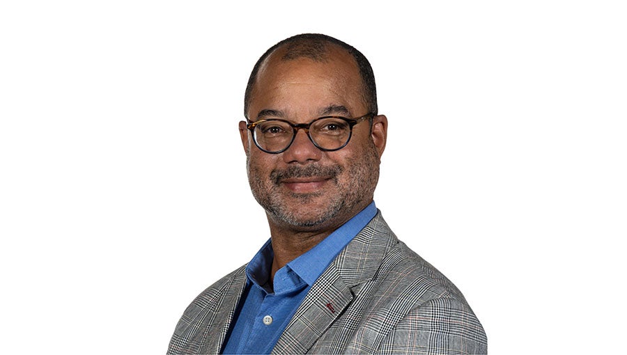 A bald man with glasses and stubble wearing a blue dress shirt and a grey suit coat.