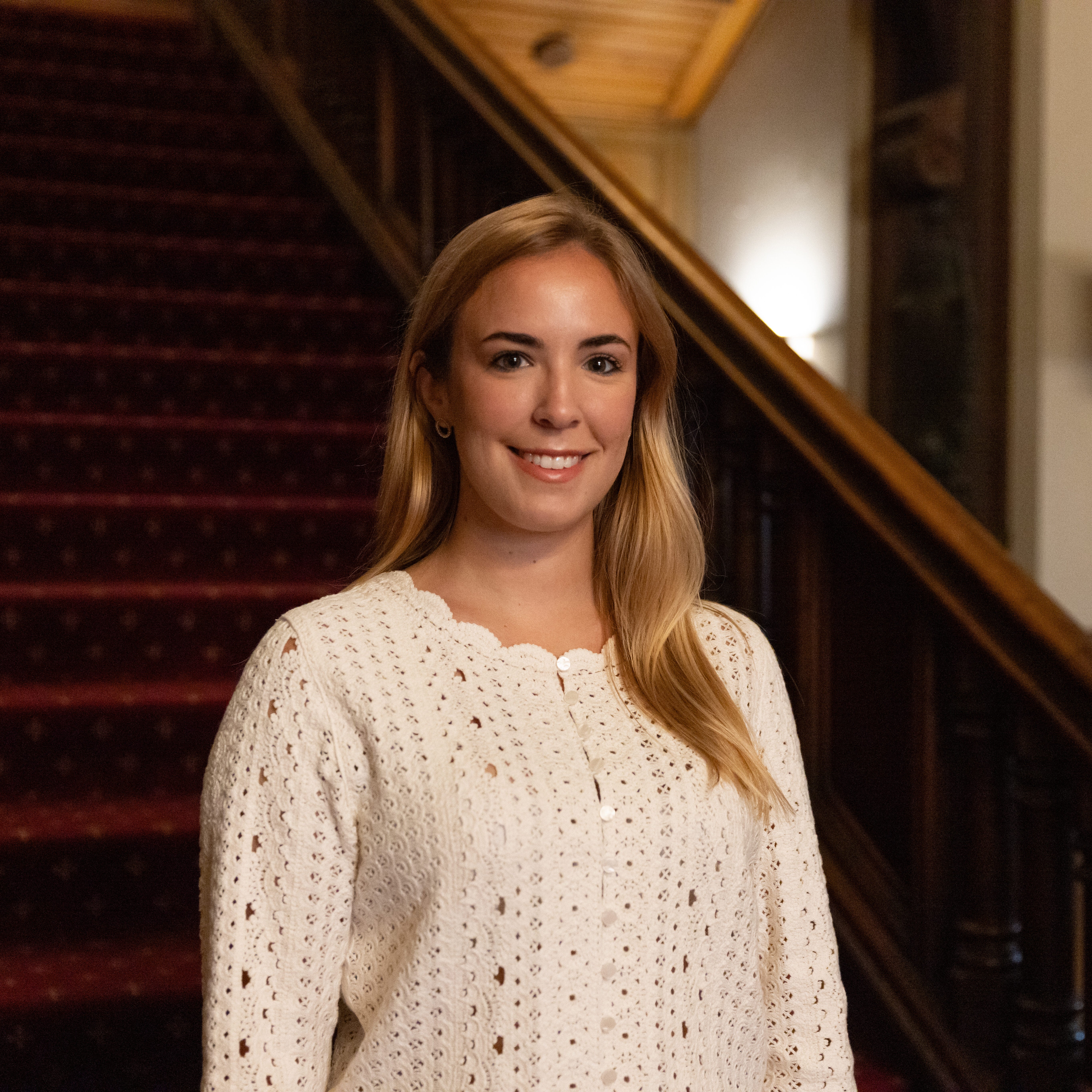 A person in a white sweater stands in front of a grand staircase with red carpet, featuring ornate wooden railings and intricate details on the side.
