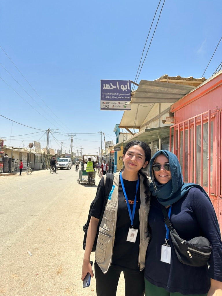 Two individuals posing alongside a busy street. Storefront facades appear in the background.