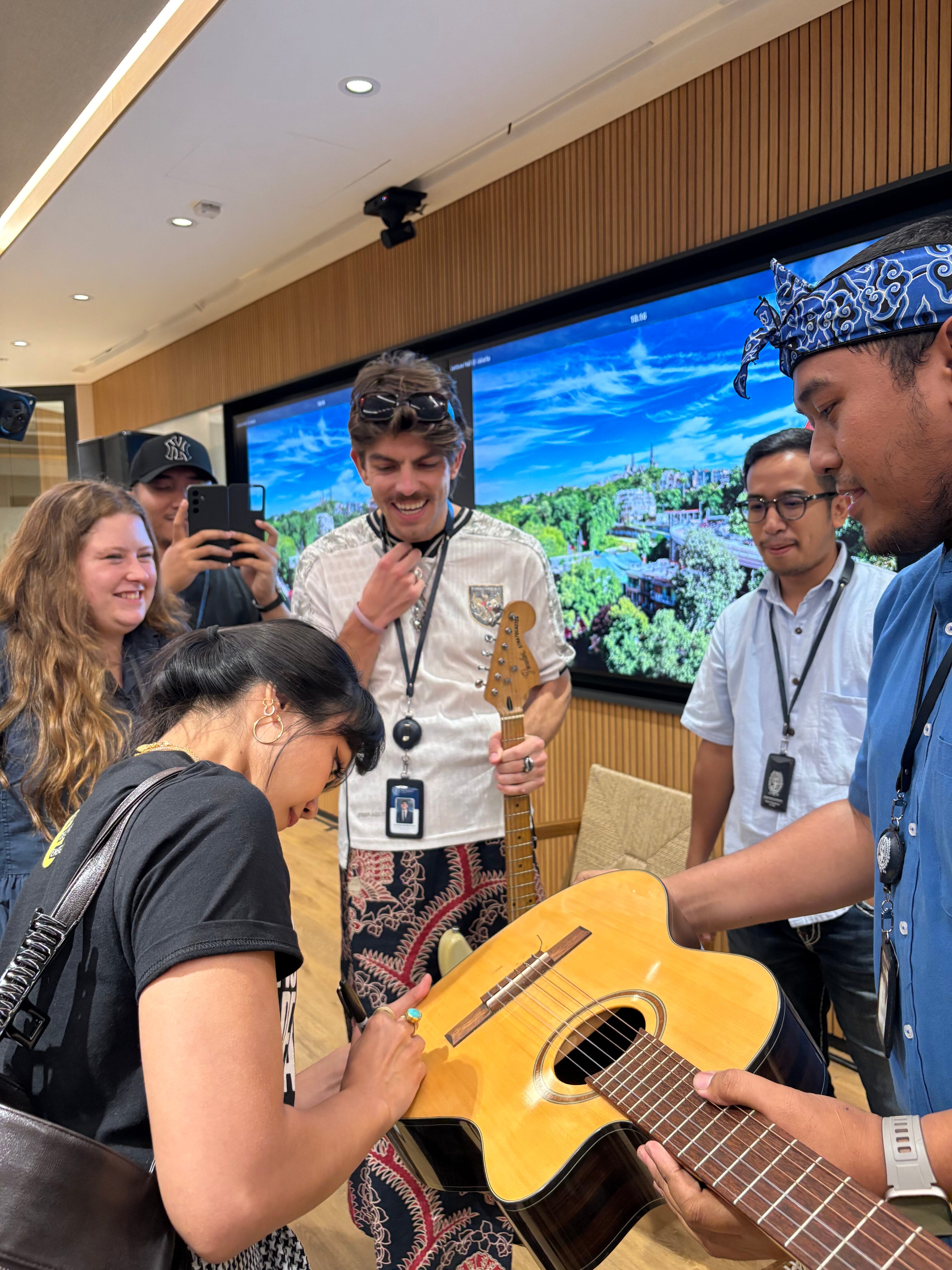 A group of people are gathered around a person signing a guitar. Two individuals hold the instruments, a guitar and a bass. Others are watching, some taking photos with phones. Behind them is a large screen displaying a scenic view.
