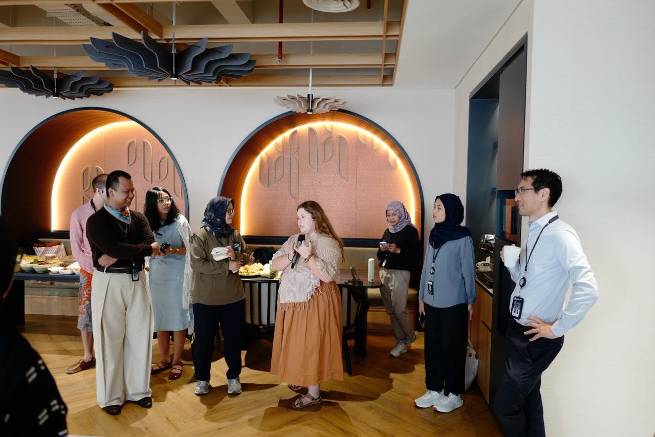 A group of people are socializing in a modern room with circular wall designs. Some individuals hold drinks, and a table with food is visible in the background. The setting appears to be casual and friendly.