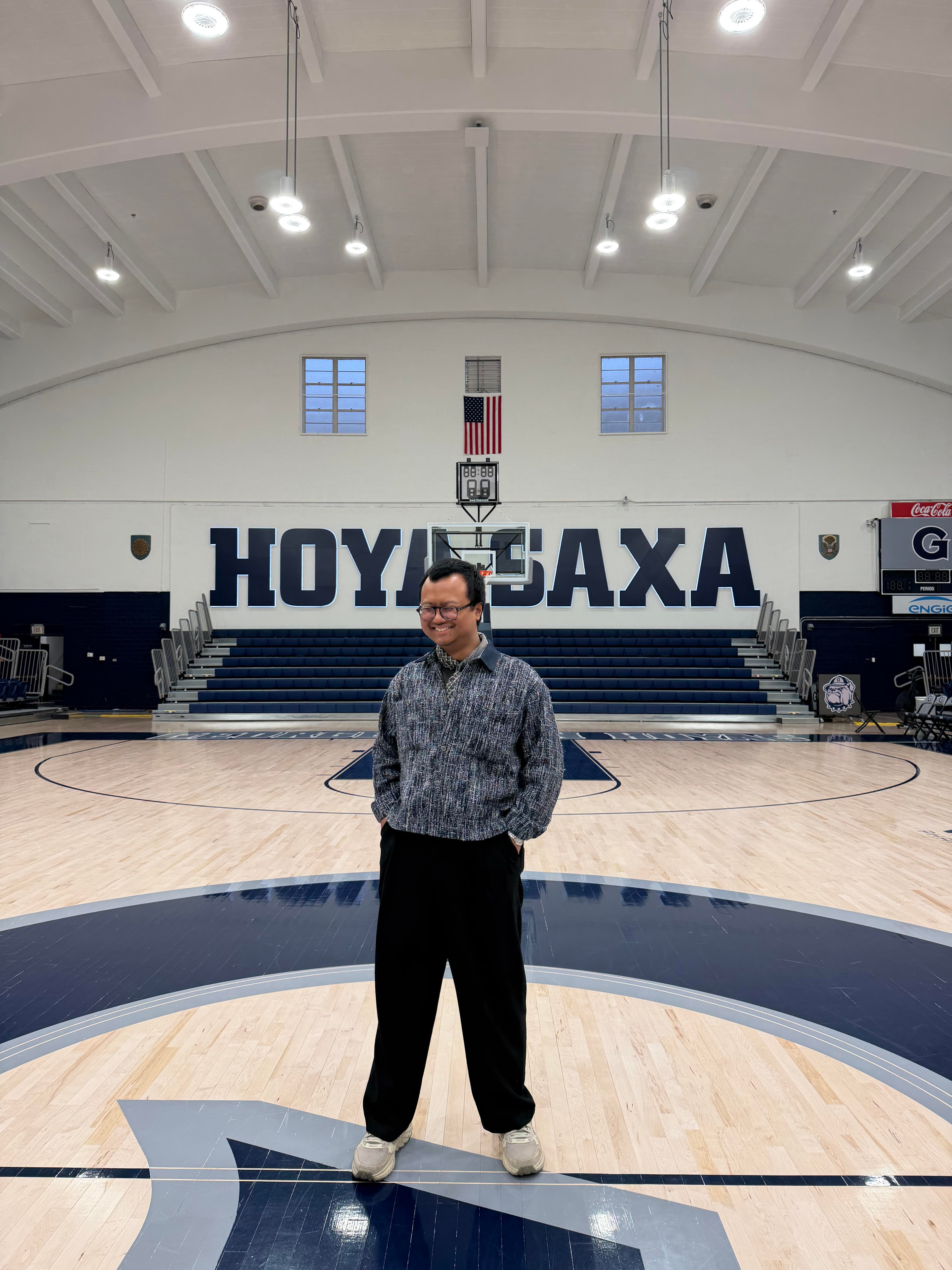 A person stands on a basketball court at Georgetown University, with "HOYA SAXA" displayed prominently on the wall behind. The court is indoors, with empty bleachers and a high ceiling.