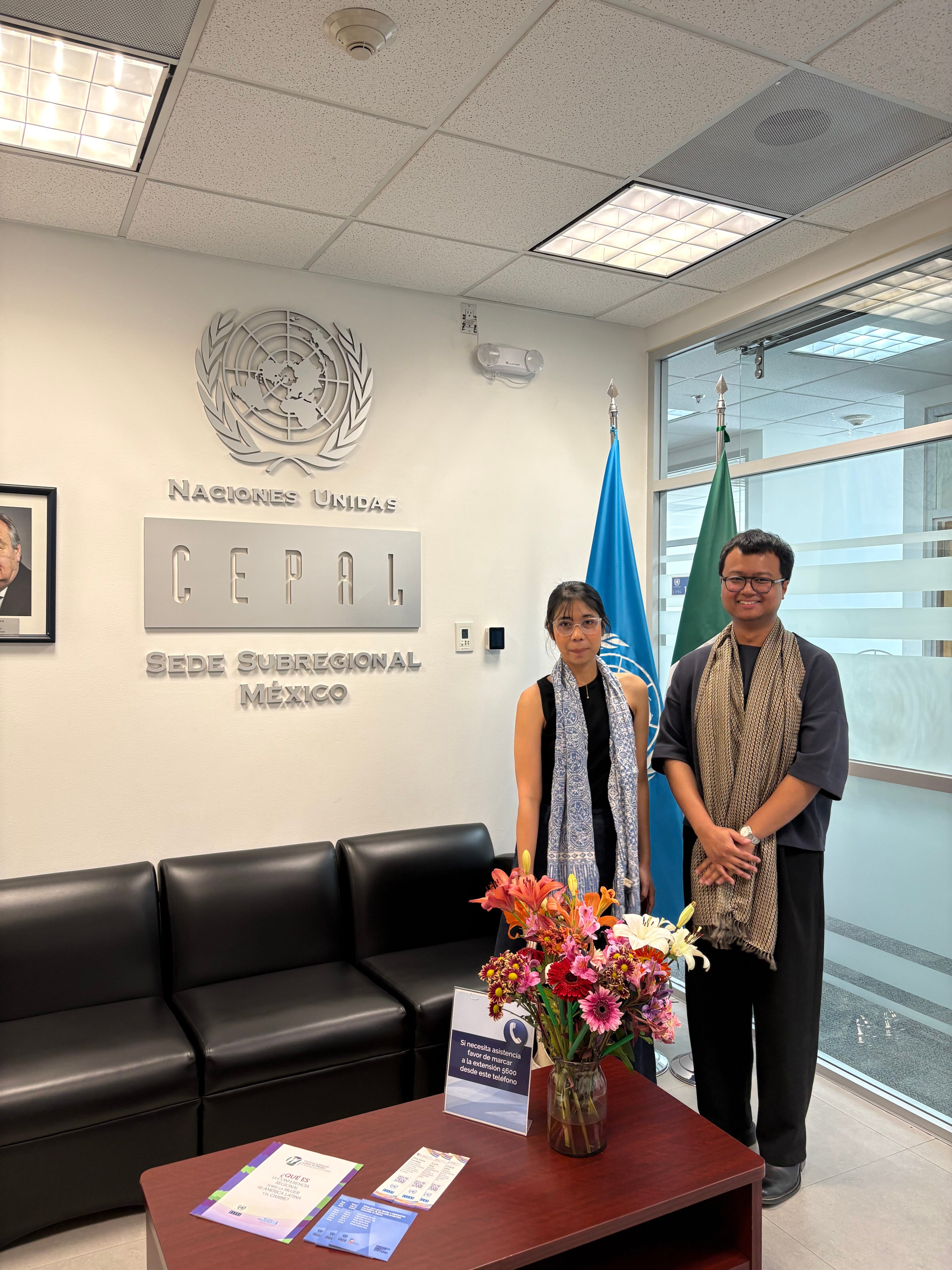 Two people stand in an office at the United Nations Economic Commission for Latin America and the Caribbean (ECLAC) Subregional Headquarters in Mexico. They are near a couch with a table displaying a bouquet of flowers and brochures.