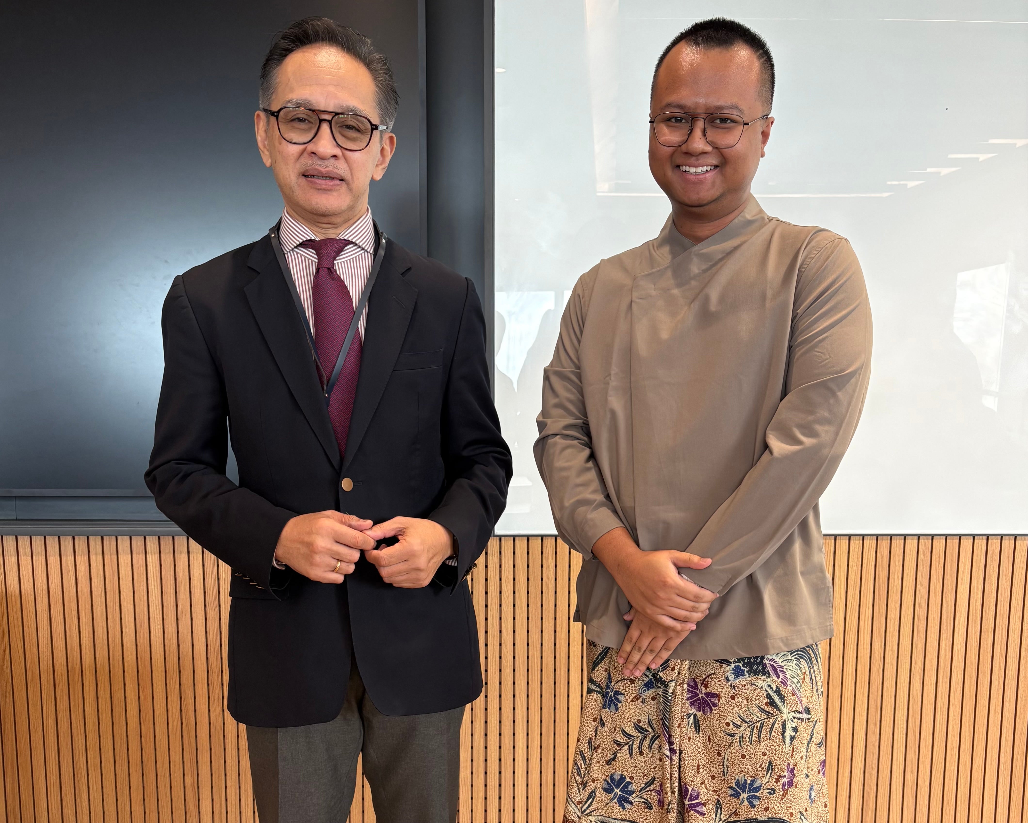 Two individuals posing in front of a whiteboard. The person on the left wears a suit with a purple tie, the person on the white wears a brown shirt and patterned pants.