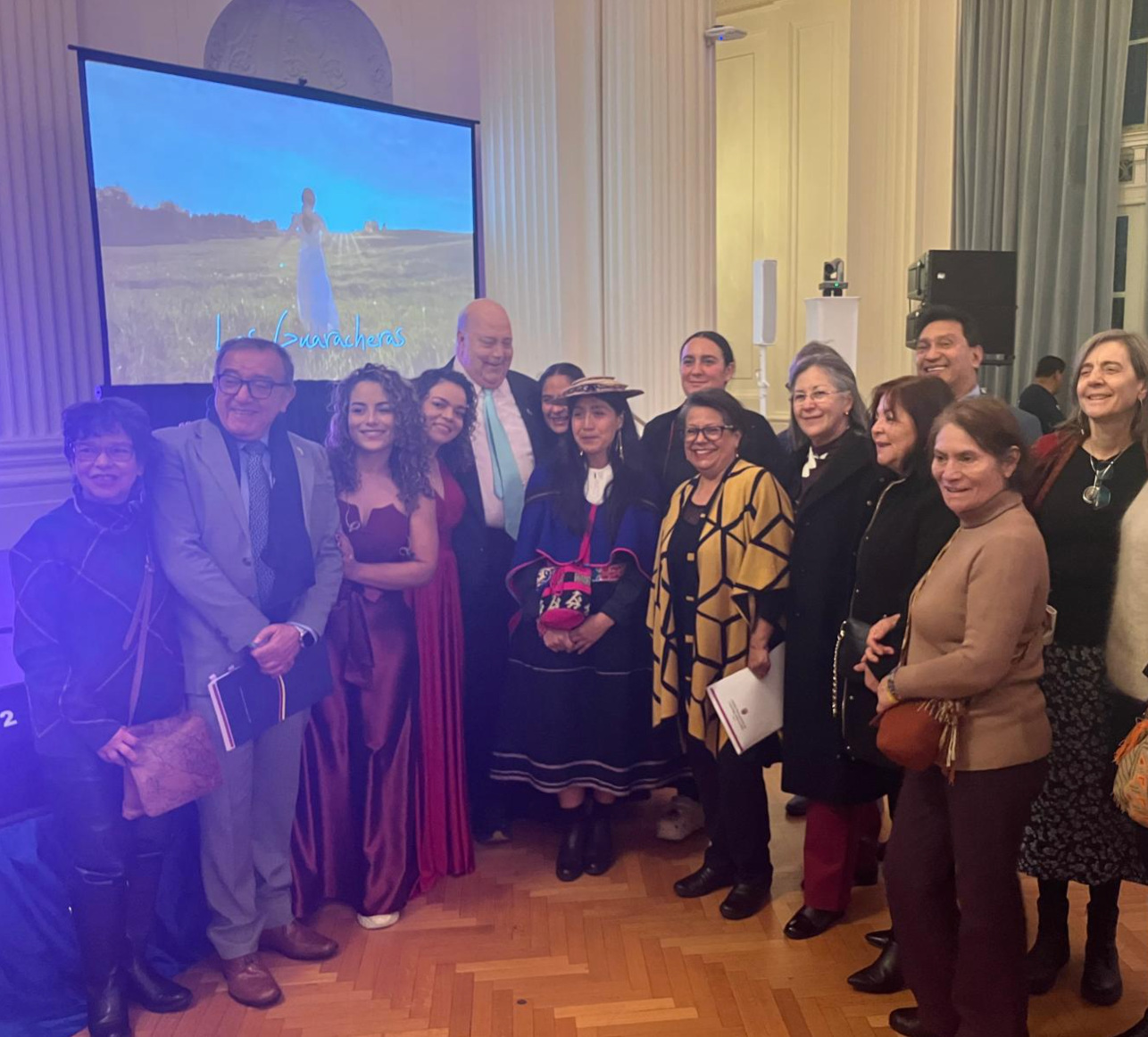 A group of 15 people in both Colombian traditional clothing and formal attire posing for a photo in front of a bright TV screen in an indoor setting.