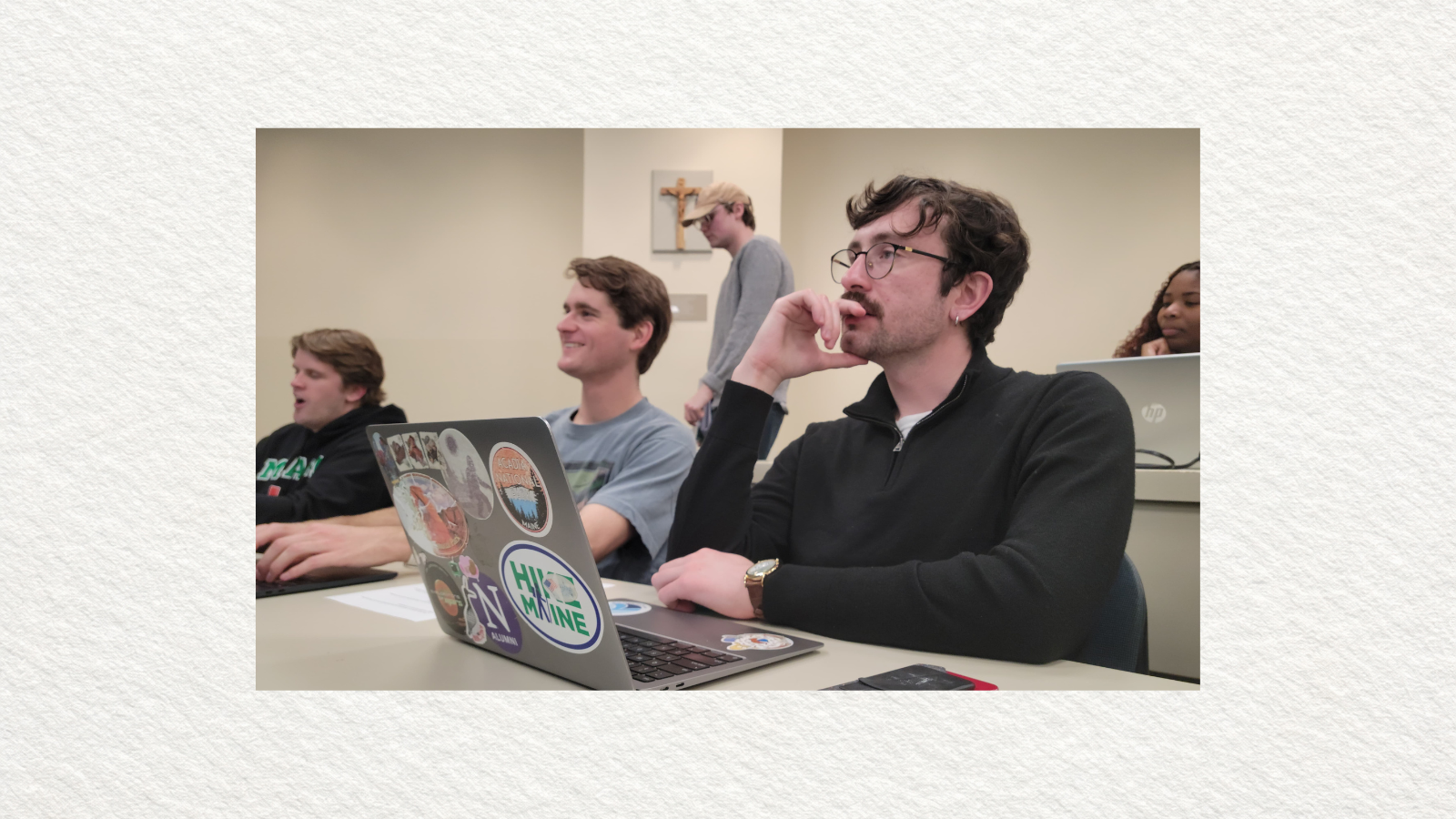 People sit classroom with laptops; one front has sticker-covered laptop while others work and person walks past a wall cross.