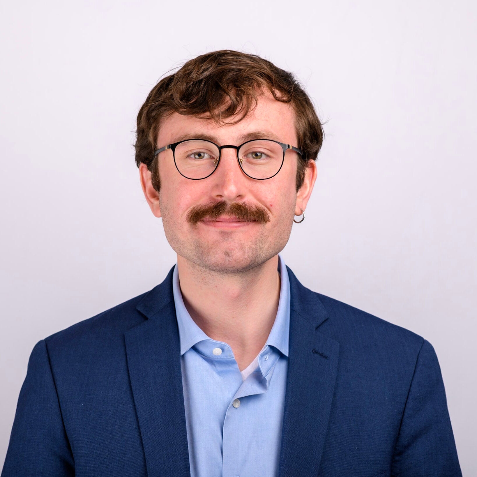 An individual with short brown hair and a mustache, wearing glasses, a blue shirt, and a navy blazer.