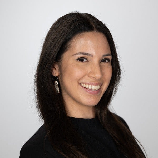 Samara Schuman dressed in black, standing in front of a grey background.