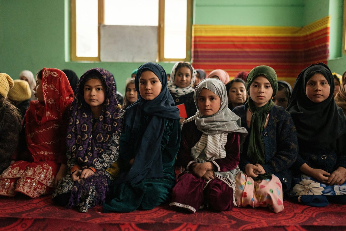 A group of children wearing colorful attire is seated indoors on a red carpet. The room has green walls and a window is in the background.