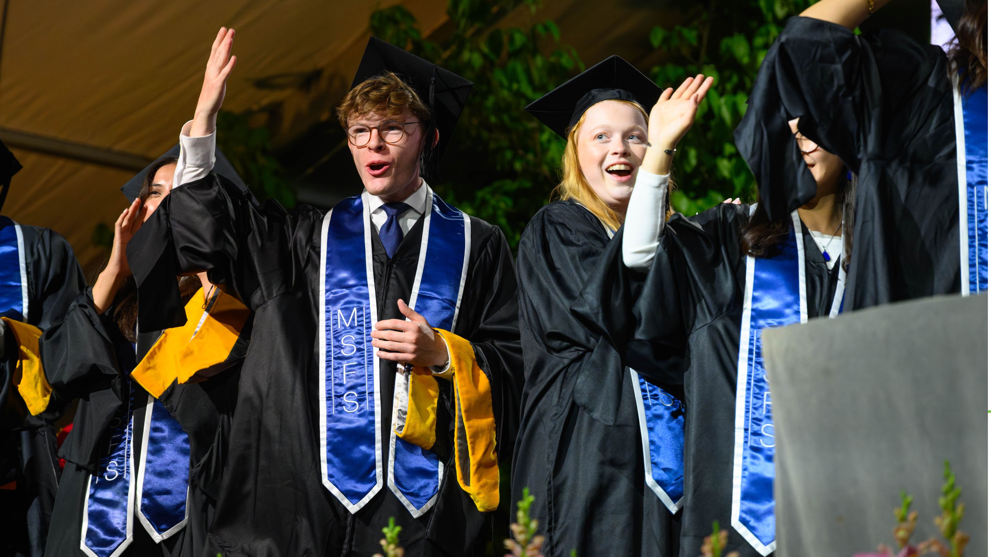 A group of people in graduation attire, including caps and gowns, are standing and gesturing with their hands raised. They are wearing blue stoles with "MSFS" written on them. The background features greenery and part of a stage.