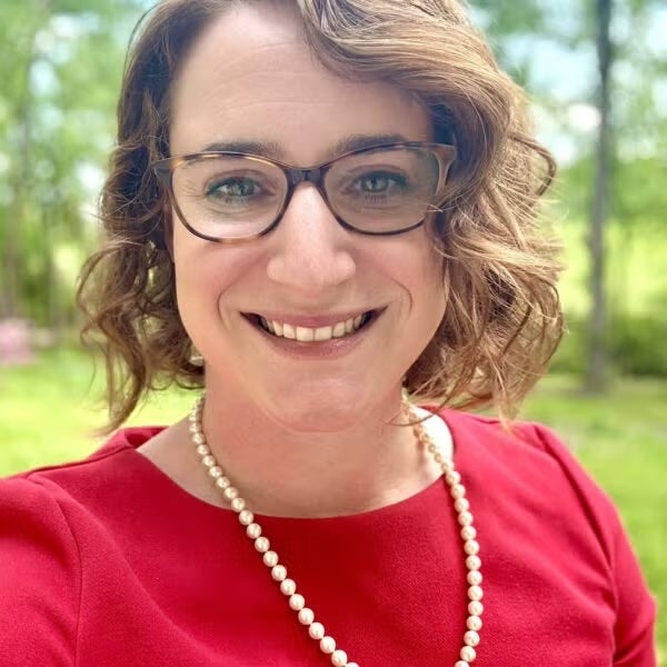 A woman with short brown hair and glasses, wearing a red top and pearl necklace.
