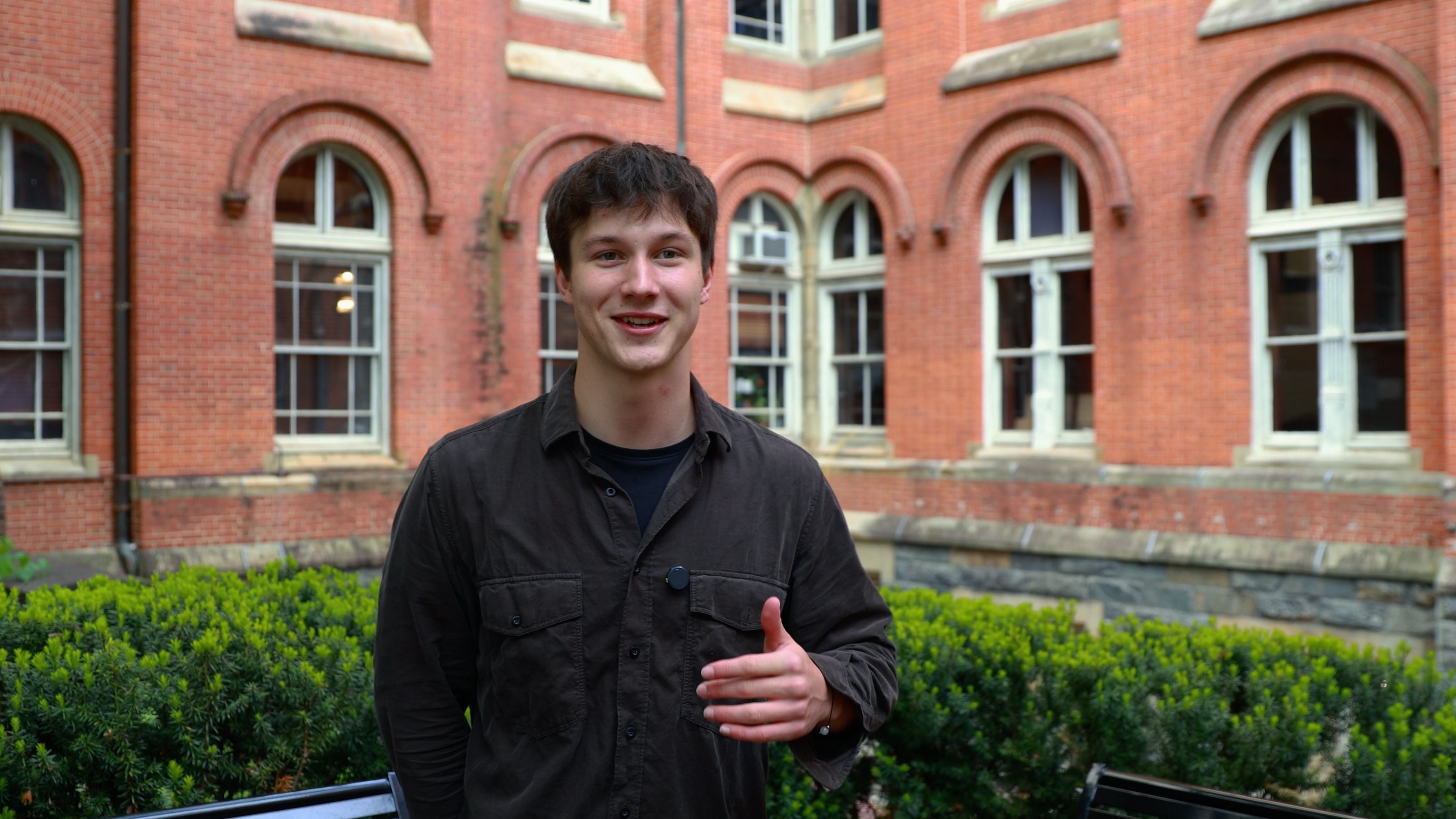 A man with short brown hair wearing a brown button up shirt speaks in front of a red brick building.