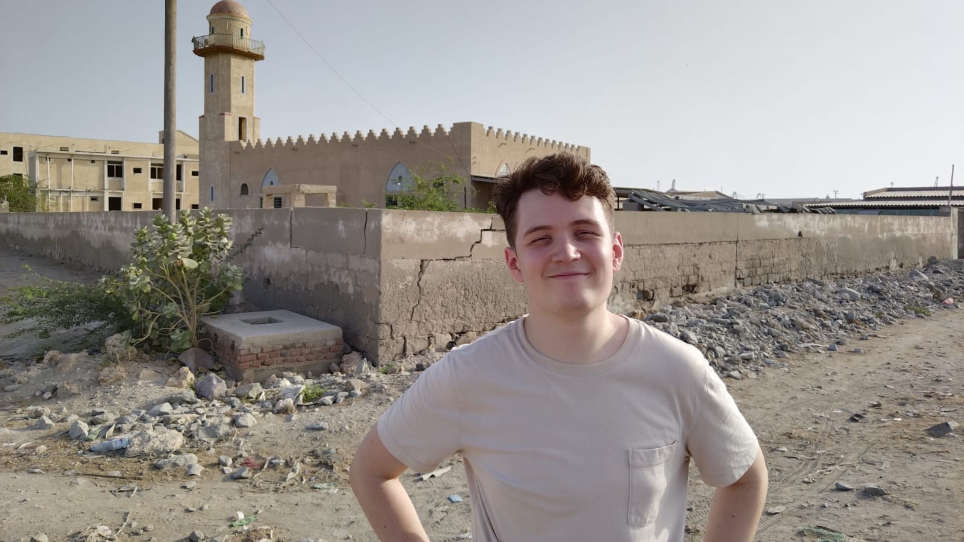 A person stands smiling in front of a low wall, with a historical building featuring a tower in the background.