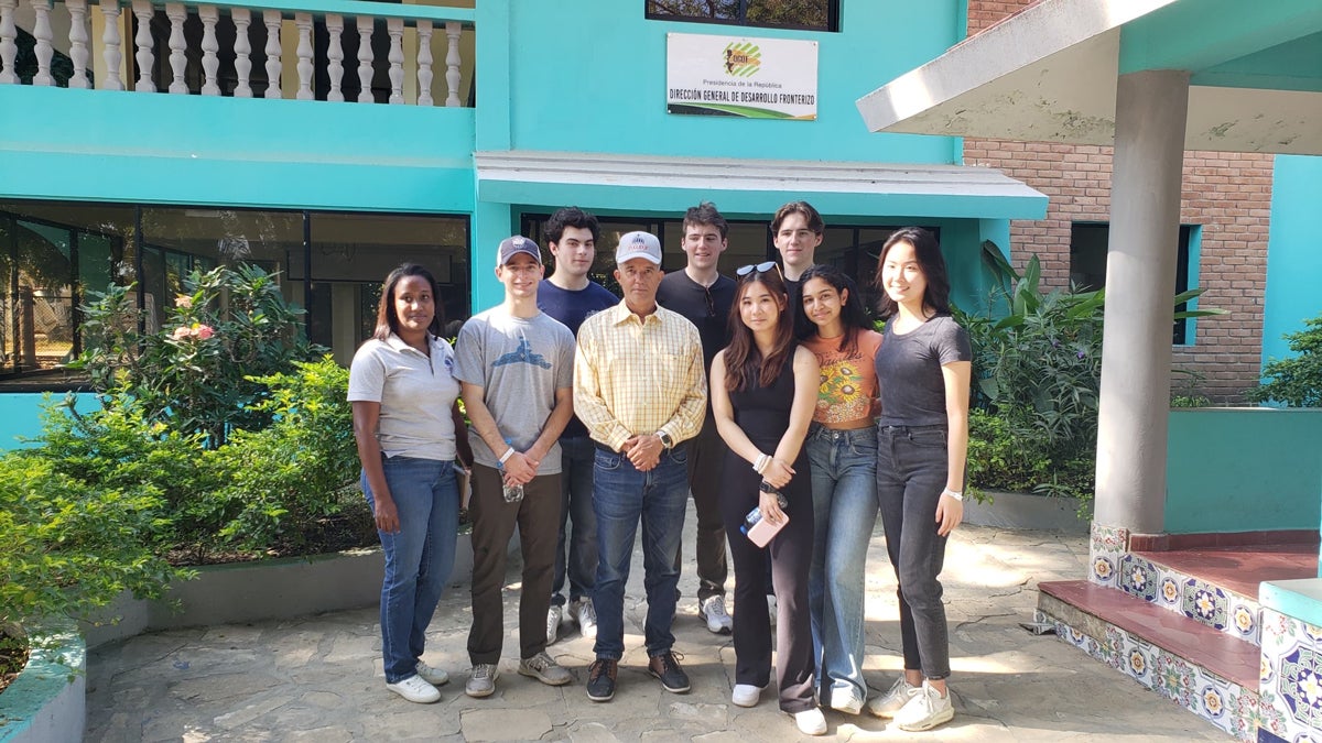 A group of eight students stand outside of a bright blue building, posing with an older man in a yellow shirt.