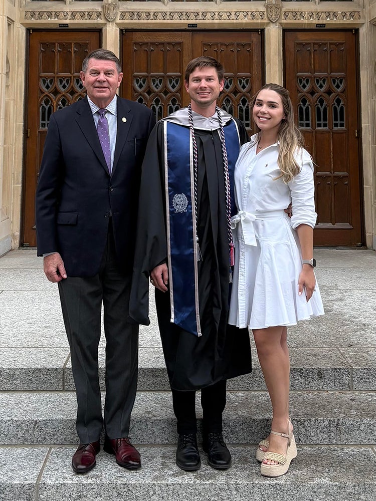 A male graduate in a gown and blue stole posing with an older man and a young woman on stone steps.