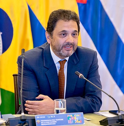 Juan Manuel Escalante sits behind a desk and microphone, colorful flags make up the background