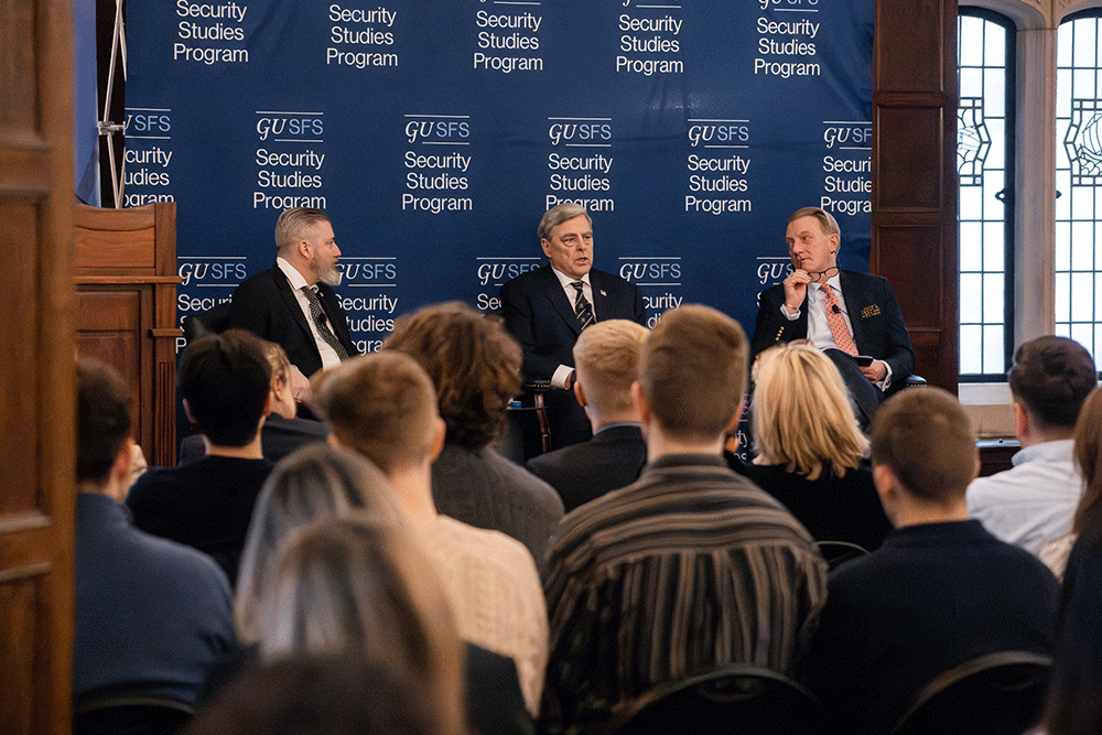 Three men speak on a panel with live audience watching