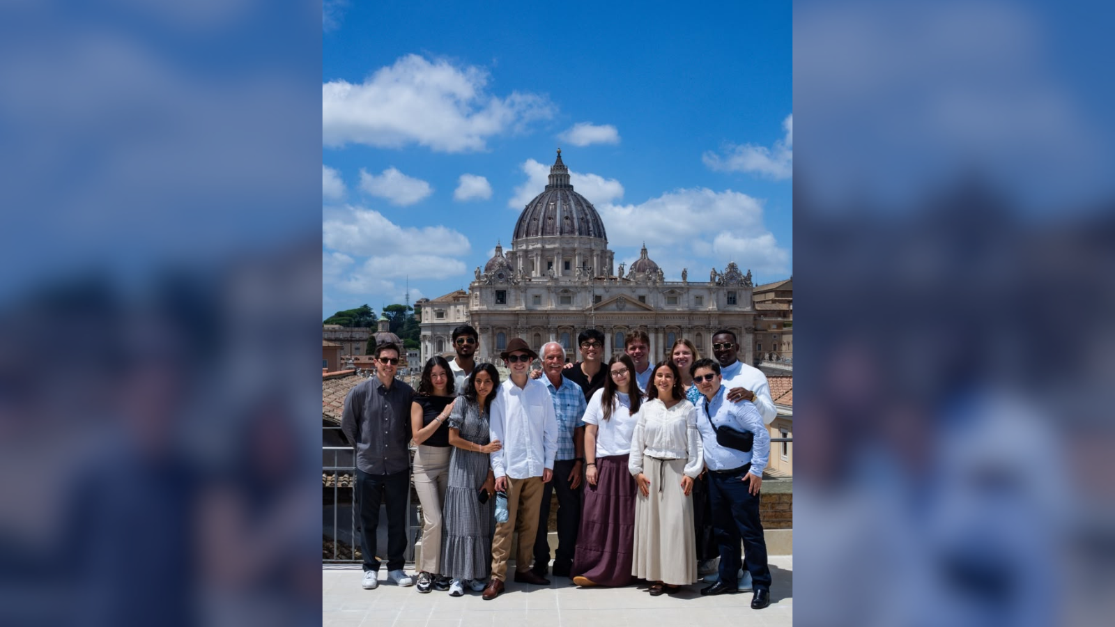 A group of people is standing together on a rooftop with St. Peter's Basilica in the background. They are smiling and dressed in a mix of casual and formal attire. The sky is clear and blue.