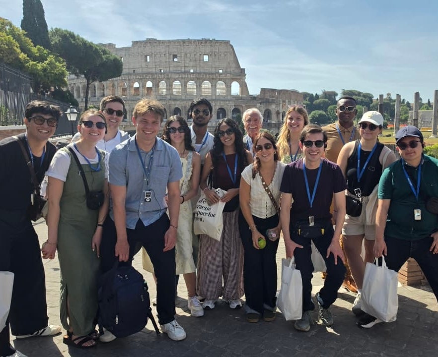 A group of tourists stands smiling in front of the Colosseum in Rome. They are wearing casual clothing and carrying small bags and cameras. The sky is clear and it appears to be daytime.