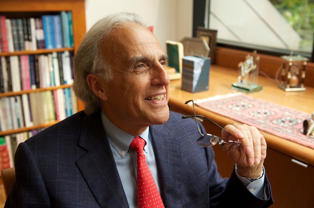 An individual is seated in an office, holding glasses with one hand and looking upwards with a smile. The person is wearing a suit with a red tie. A bookshelf with various books is visible in the background, alongside a wooden desk with decorative items.