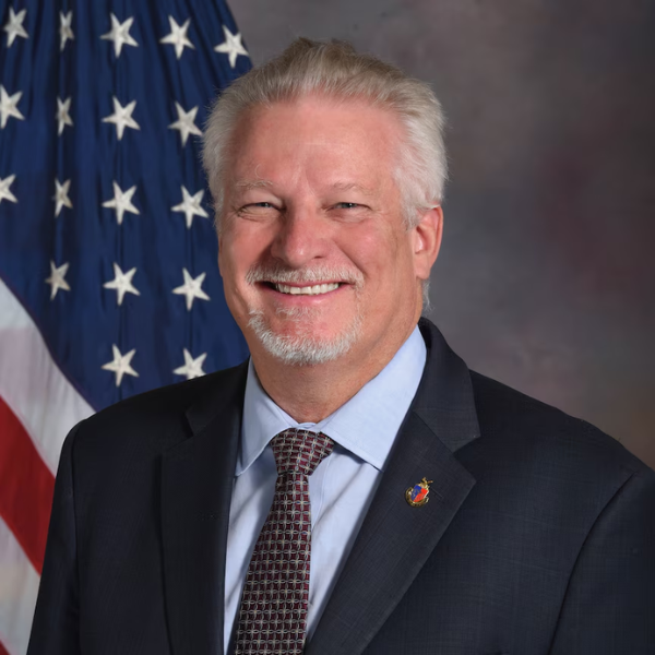 An individual with short white hair, wearing a black suit and tie in front of an American flag.