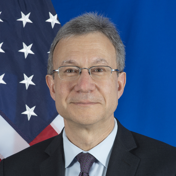An individual wearing glasses, a black suit, and tie in front of an American flag.