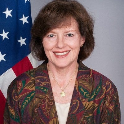 An individual with short brown hair wearing a brown, multi-colored blouse in front of an American flag.