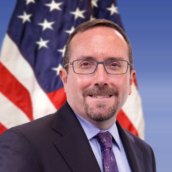 A person wearing a suit, tie, and glasses poses in front of an American flag.