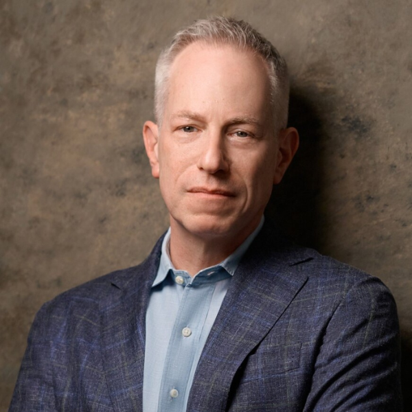 An individual wearing a gray suit and blue shirt standing in front of a brown background.