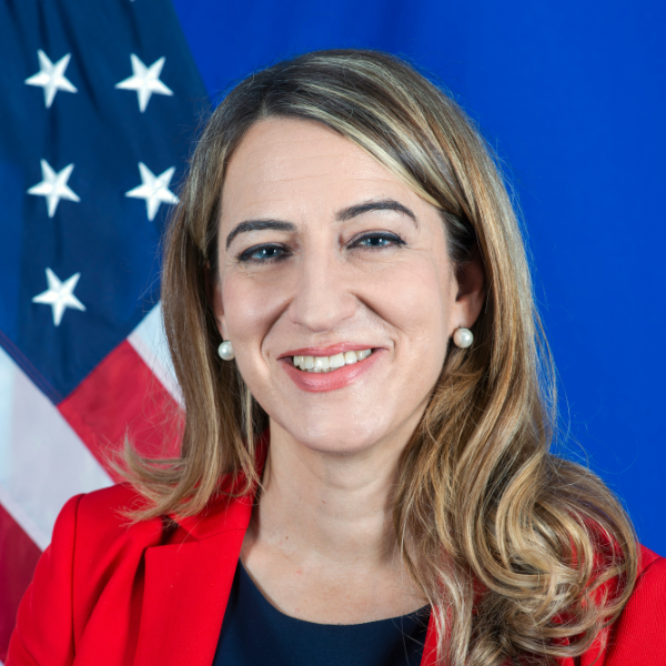 An individual with long blonde hair wearing a red blazer in front of an American flag.
