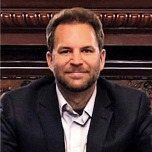 An individual wearing a black blazer and white shirt in front of a brown wooden background.