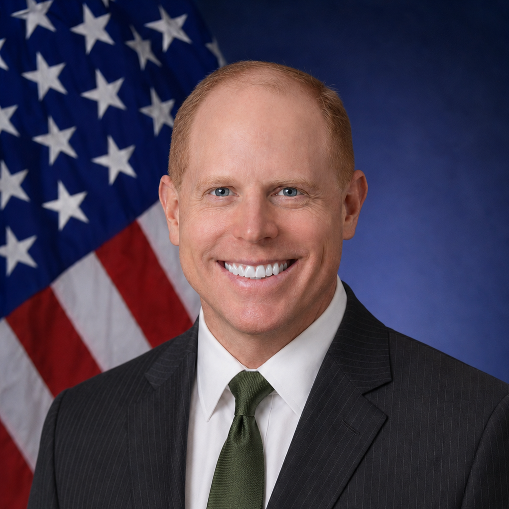An individual wearing a black blazer and gray tie in front of an American flag.